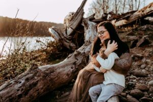 mom holds son and smiles with river and drift wood in the background